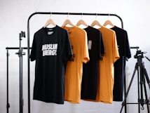 Four t-shirts, two black and two yellow, are hanging on wooden hangers from a black metal clothes rack. The shirts have different texts printed on them. There is a camera tripod on the right side of the frame. The background is a white brick wall.
