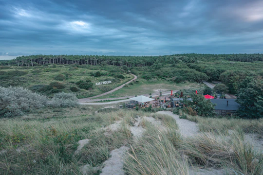 A grassy, sandy landscape with a path leading to a small building surrounded by trees. A large sign reads 'KAAP HOORN' on the hillside. The scene is under a cloudy sky, creating a tranquil, serene atmosphere.