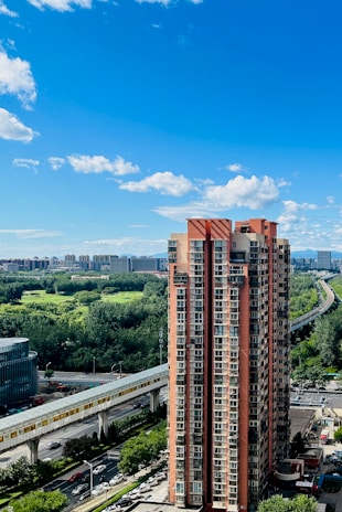 A tall red and beige residential building stands prominently in the foreground, with lush green trees, a park or open green space, and several other buildings visible in the background under a clear blue sky. A raised train track runs parallel to the scene, bordered by roads with visible vehicles.