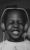 A child wearing large headphones smiles broadly with eyes closed, exuding a joyful and carefree expression. The image is in black and white, emphasizing the child's happy demeanor.