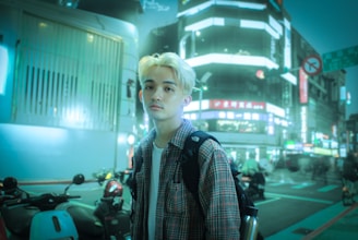 A young person with light blonde hair and casual attire stands on a city street at night. They carry a backpack and are surrounded by numerous motorbikes. Bright, neon signs illuminate the background, contributing to a bustling urban atmosphere.