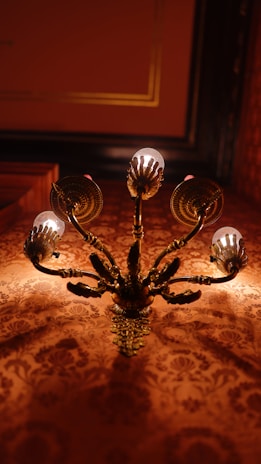An ornate chandelier with golden bronze detailing and multiple arms supports small light bulbs. The intricate wall behind it features a richly patterned design in warm tones, contributing to an overall luxurious and classic ambiance.