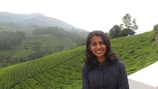 A person smiling in the foreground with a background of lush green hills covered in tea plantations. The landscape is mountainous and partially covered with mist, creating a serene and peaceful atmosphere.