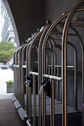 A row of empty metallic luggage carts are lined up neatly against a dark wall. These carts are typically used in hotels to transport guests' luggage. The background features part of an urban setting with buildings and trees visible at a distance. The scene is illuminated by natural light.