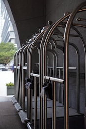A row of empty metallic luggage carts are lined up neatly against a dark wall. These carts are typically used in hotels to transport guests' luggage. The background features part of an urban setting with buildings and trees visible at a distance. The scene is illuminated by natural light.