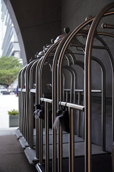 A row of empty metallic luggage carts are lined up neatly against a dark wall. These carts are typically used in hotels to transport guests' luggage. The background features part of an urban setting with buildings and trees visible at a distance. The scene is illuminated by natural light.