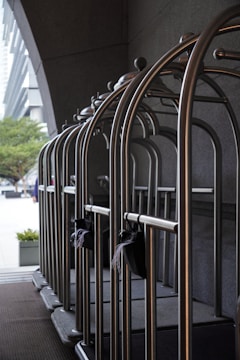 A row of empty metallic luggage carts are lined up neatly against a dark wall. These carts are typically used in hotels to transport guests' luggage. The background features part of an urban setting with buildings and trees visible at a distance. The scene is illuminated by natural light.