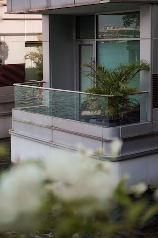 A modern glass balcony extends from a building, surrounded by lush green plants. The reflection of water can be seen below, creating a serene waterfront setting. The building has a contemporary design, with clean lines and a mix of grey and blue tones.