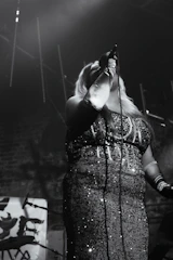 A person is performing on stage with a microphone in their hand. They are wearing a sparkling, sequined dress and multiple bracelets on their arm. The stage lighting creates a dramatic effect with shadows and highlights, and there is equipment visible in the background, including a microphone stand.