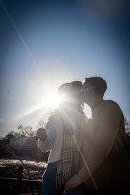 A couple shares an intimate moment, with one person leaning in to kiss the other. The sun shines brightly from behind them, casting a brilliant halo around their silhouettes. Both individuals are casually dressed, and one is wearing a scarf. In the background, there are trees and a building, suggesting an outdoor setting.
