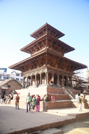 A multi-tiered, traditional wooden pagoda structure with intricate carvings stands prominently in a public square. Several people are sitting and standing around the structure, engaged in conversations and leisure activities. The surrounding area has brick pavement and there's visible scaffolding, indicating possible restoration work on nearby buildings.