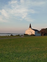 A white church with a tall black steeple is situated near a body of water. It is surrounded by a well-maintained green lawn, a paved walkway, and some low shrubs. The sky above is partly cloudy and the lighting suggests it's either early morning or late evening.
