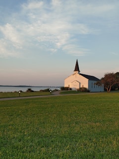 A white church with a tall black steeple is situated near a body of water. It is surrounded by a well-maintained green lawn, a paved walkway, and some low shrubs. The sky above is partly cloudy and the lighting suggests it's either early morning or late evening.