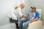 A healthcare professional wearing a white coat and gloves is attending to a man who is seated in a recliner chair. An intravenous drip is visible, indicating that the man might be receiving a medical treatment. The man is wearing a colorful tie-dye t-shirt featuring a graphic and a cap. The setting appears to be a clinical environment with minimal decor and medical equipment.
