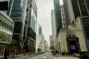 A cityscape featuring skyscrapers with reflective glass facades and modern architectural designs. The street is lined with tall buildings and has visible traffic lights and a few pedestrians walking. The weather appears overcast, suggesting a cloudy sky.
