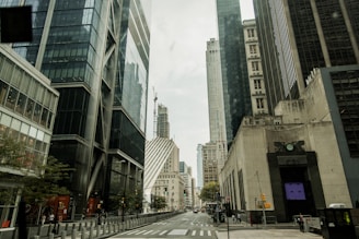 A cityscape featuring skyscrapers with reflective glass facades and modern architectural designs. The street is lined with tall buildings and has visible traffic lights and a few pedestrians walking. The weather appears overcast, suggesting a cloudy sky.
