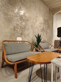 A cozy seating area with wooden benches featuring grey cushions is arranged against a textured, rustic wall. A small wooden table with a round tabletop and metal legs sits nearby. The setting includes a potted plant and a small section of a speaker or column visible in the background.