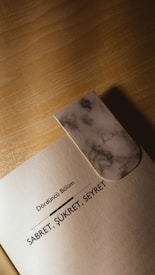 A book page with a marble-patterned bookmark resting on it. The page is titled 'D&ouml;rd&uuml;nc&uuml; B&ouml;l&uuml;m' and features the text 'Sabret, Ş&uuml;kret, Seyret' in bold letters. The bookmark is rectangular with soft edges and the word 'Sabır' is written on it in elegant script.