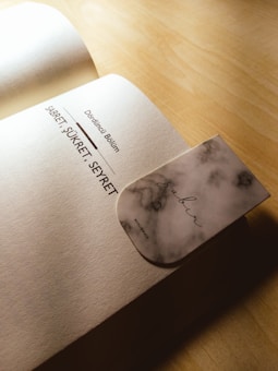 An open book on a wooden surface with a focus on a page displaying text in a non-English language. A marble-patterned bookmark with the word 'Sabr' printed on it is partially inserted into the book.