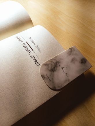 An open book on a wooden surface with a focus on a page displaying text in a non-English language. A marble-patterned bookmark with the word 'Sabr' printed on it is partially inserted into the book.