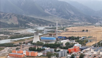 An industrial facility with a large cooling tower and smokestack is situated in a valley surrounded by mountains. There is a river running alongside the facility, bordered by fields and various buildings, including some with orange and blue roofs.