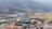 An industrial facility with a large cooling tower and smokestack is situated in a valley surrounded by mountains. There is a river running alongside the facility, bordered by fields and various buildings, including some with orange and blue roofs.