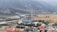 An industrial facility with a large cooling tower and smokestack is situated in a valley surrounded by mountains. There is a river running alongside the facility, bordered by fields and various buildings, including some with orange and blue roofs.