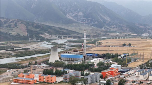 An industrial facility with a large cooling tower and smokestack is situated in a valley surrounded by mountains. There is a river running alongside the facility, bordered by fields and various buildings, including some with orange and blue roofs.