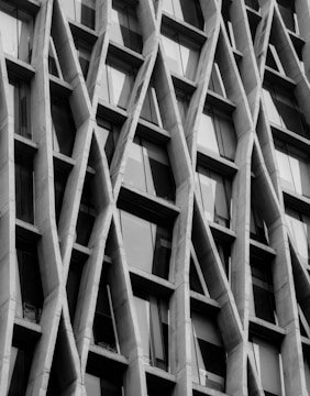 A close-up view of a tall building's exterior features diagonal concrete beams that intersect in a lattice-like pattern. The structure has numerous windows, likely glass, set within this geometric framework, creating a modern architectural appearance.