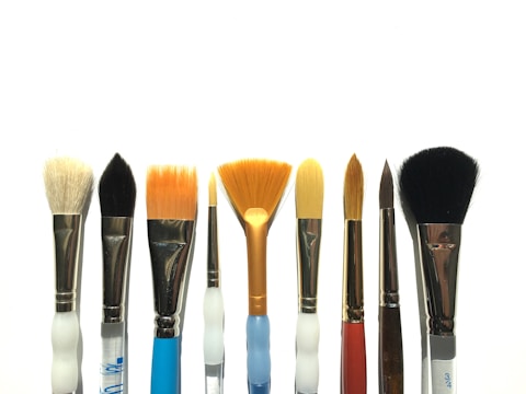 A collection of paintbrushes with a variety of shapes and sizes arranged in a row against a white background. The bristles range from flat and fan-shaped to round tips, and they appear to be made from different materials.