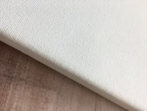 A close-up view of the corner of a blank, textured canvas resting on a wooden surface. The canvas is slightly tilted and the texture is visibly detailed.