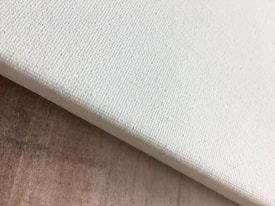 A close-up view of the corner of a blank, textured canvas resting on a wooden surface. The canvas is slightly tilted and the texture is visibly detailed.