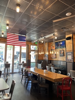 The interior features wooden paneling and metal detailing with industrial-style light fixtures hanging from the ceiling. An American flag is displayed prominently near the ceiling. There are tables and chairs arranged in a casual dining setting, and a television mounted on the wall showing a program. Natural light streams in through large windows, creating a bright and inviting atmosphere.