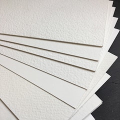 A stack of textured white sheets of paper arranged in a fanned-out manner on a dark surface. The paper has a subtle grainy texture, and each sheet seems thick and sturdy.