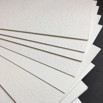A stack of textured white sheets of paper arranged in a fanned-out manner on a dark surface. The paper has a subtle grainy texture, and each sheet seems thick and sturdy.