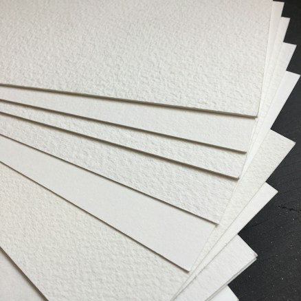 A stack of textured white sheets of paper arranged in a fanned-out manner on a dark surface. The paper has a subtle grainy texture, and each sheet seems thick and sturdy.