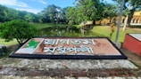A large sign with text in a non-Latin script is prominently displayed. The sign features a map embedded in it and is situated by the edge of a pond surrounded by lush green trees. A pathway can be seen leading into the distance, with a building partially visible on the right.