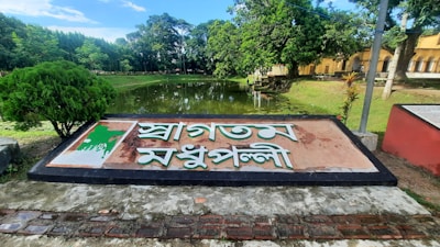 A large sign with text in a non-Latin script is prominently displayed. The sign features a map embedded in it and is situated by the edge of a pond surrounded by lush green trees. A pathway can be seen leading into the distance, with a building partially visible on the right.