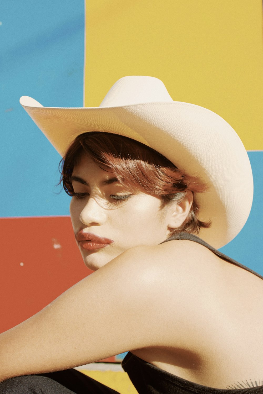 a woman wearing a white cowboy hat sitting against a colorful wall