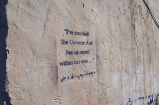 A textured concrete wall features a piece of graffiti with stencil-style text in both English and another language. The poem expresses introspection and connection, with a focus on finding oneself in another's eyes.