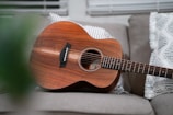An acoustic guitar with a rich wooden finish is resting on a sofa, surrounded by patterned throw pillows. The scene is well-lit, with soft natural light coming through window blinds in the background.