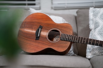 An acoustic guitar with a rich wooden finish is resting on a sofa, surrounded by patterned throw pillows. The scene is well-lit, with soft natural light coming through window blinds in the background.