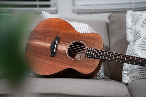 An acoustic guitar with a rich wooden finish is resting on a sofa, surrounded by patterned throw pillows. The scene is well-lit, with soft natural light coming through window blinds in the background.