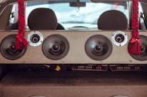 The image is taken from inside a vehicle, looking towards the rear with a focus on a large speaker system mounted in the back. The speaker setup includes multiple round audio speakers and tweeters arranged across a panel. Two red and black braided tassels hang from the ceiling on either side of the speakers, adding a decorative element.