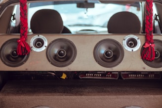 The image is taken from inside a vehicle, looking towards the rear with a focus on a large speaker system mounted in the back. The speaker setup includes multiple round audio speakers and tweeters arranged across a panel. Two red and black braided tassels hang from the ceiling on either side of the speakers, adding a decorative element.