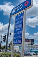 A blue and white gas station sign with 'ARCO' at the top and prices listed for different types of gasoline both for cash and credit payments. In the background, a billboard advertises a TV show above buildings and cars on a busy street. Trees and a traffic light are also visible.