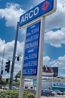 A blue and white gas station sign with 'ARCO' at the top and prices listed for different types of gasoline both for cash and credit payments. In the background, a billboard advertises a TV show above buildings and cars on a busy street. Trees and a traffic light are also visible.