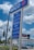 A blue and white gas station sign with 'ARCO' at the top and prices listed for different types of gasoline both for cash and credit payments. In the background, a billboard advertises a TV show above buildings and cars on a busy street. Trees and a traffic light are also visible.