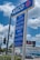 A blue and white gas station sign with 'ARCO' at the top and prices listed for different types of gasoline both for cash and credit payments. In the background, a billboard advertises a TV show above buildings and cars on a busy street. Trees and a traffic light are also visible.