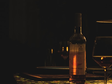 A dimly lit scene featuring a bottle of Merlot Rosé wine on a tablecloth with geometric patterns. The bottle's label is partially illuminated, casting a warm glow. There are glasses partially filled with wine set around, creating an intimate and cozy ambiance.
